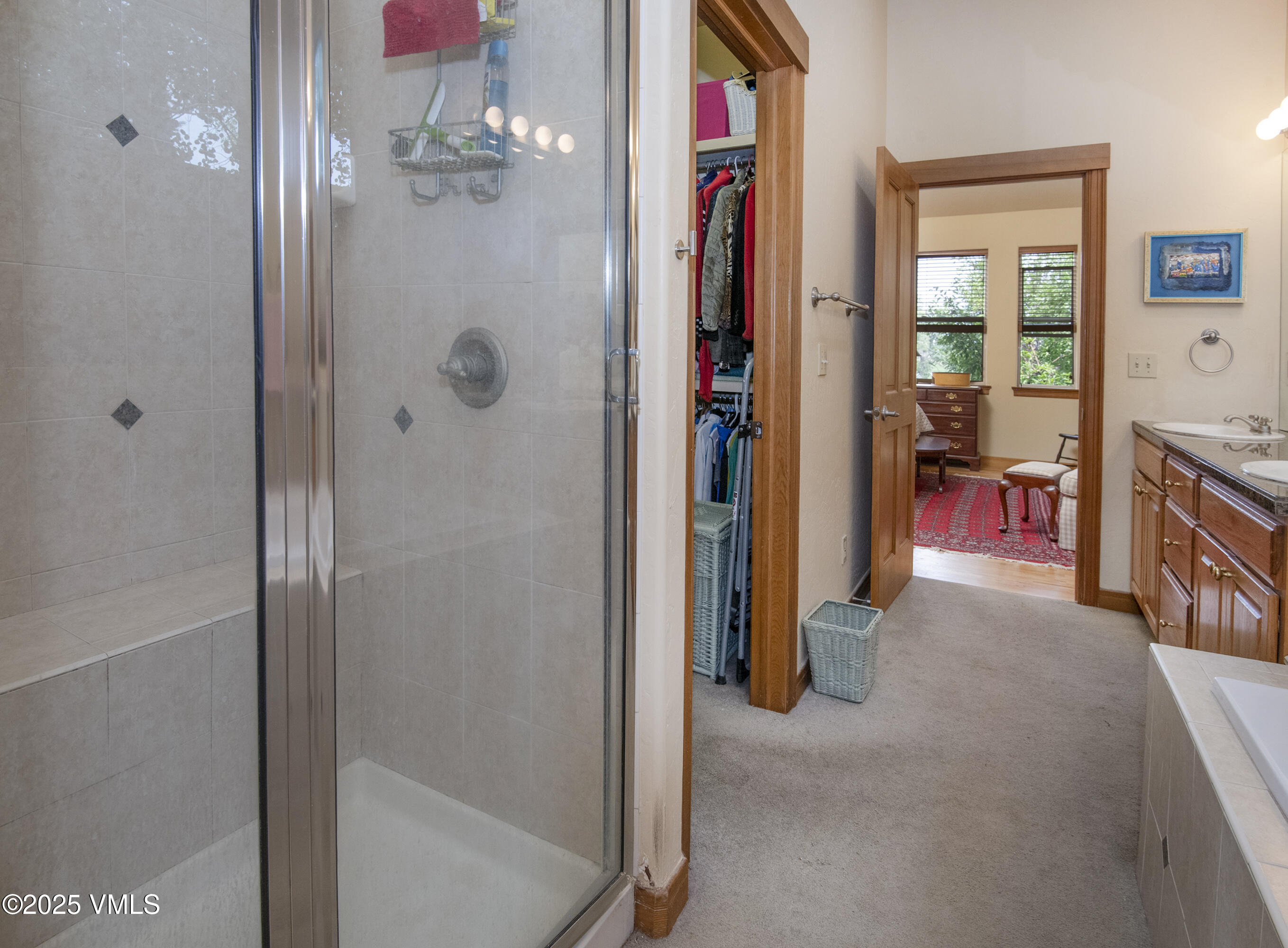 Bathroom with a glass-enclosed tiled shower, double wood vanity, and open closet leading to a sitting area.