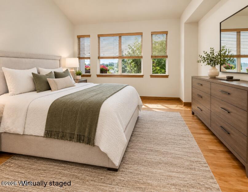 Staged bedroom with a large bed, wide windows, wooden dresser, and hardwood flooring.