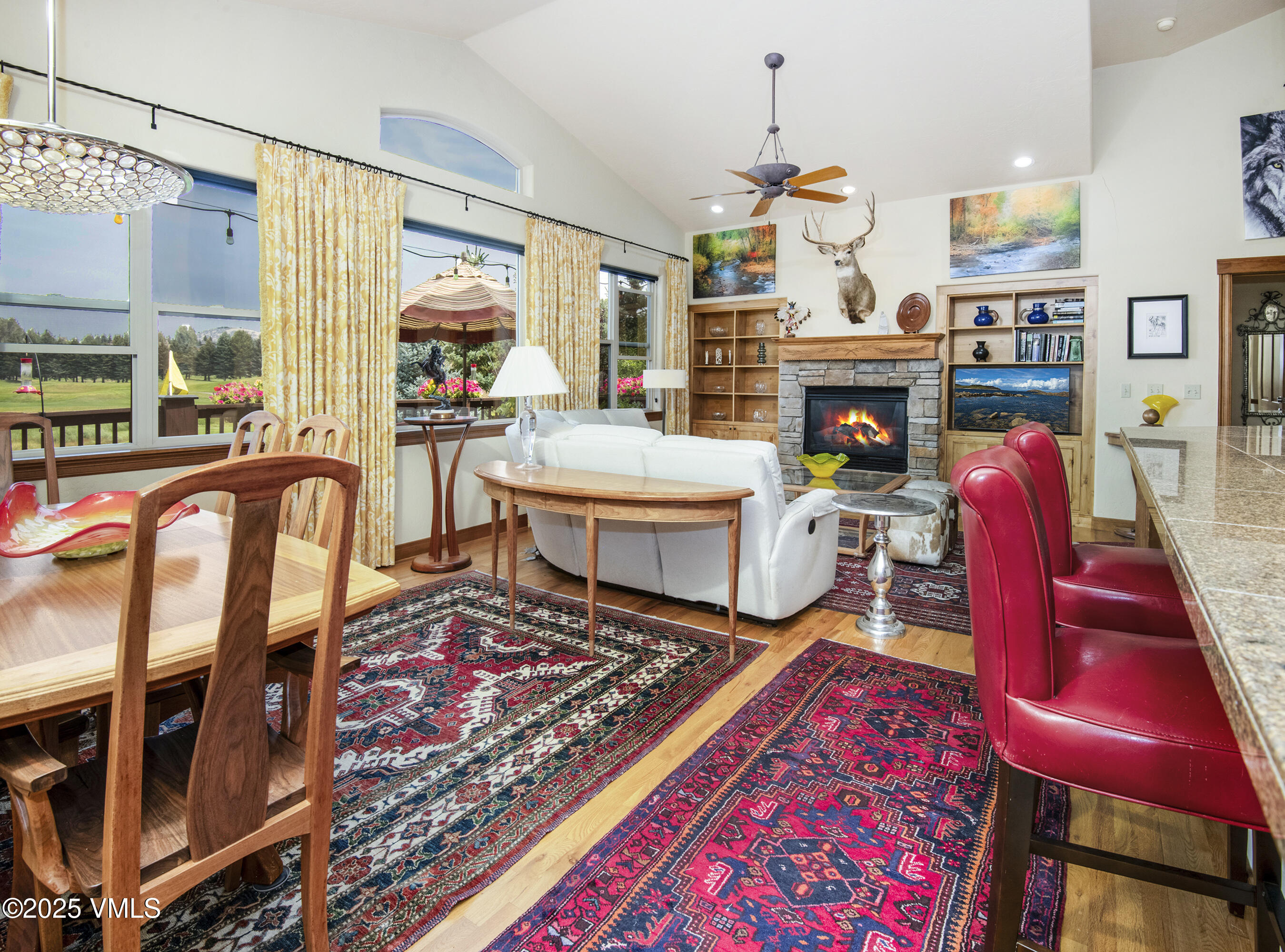 Open living and dining room with vaulted ceiling, stone fireplace, and large windows overlooking a lawn.