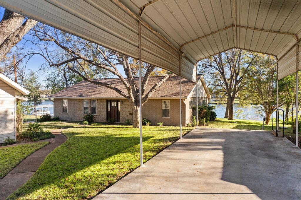 Lakefront house viewed from beneath a covered carport, with a paved driveway and mature trees.