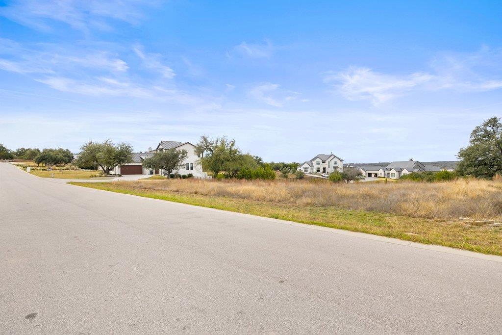 Open grassy lot beside a paved road, with neighboring homes and low hills in the distance.