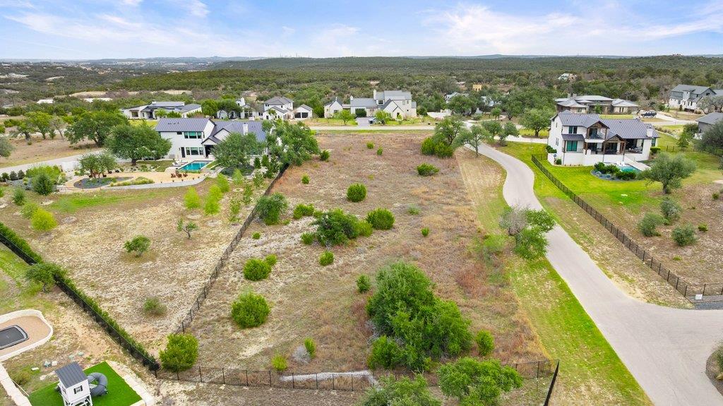 Vacant fenced lot beside a paved road, with neighboring homes and wooded hills beyond.