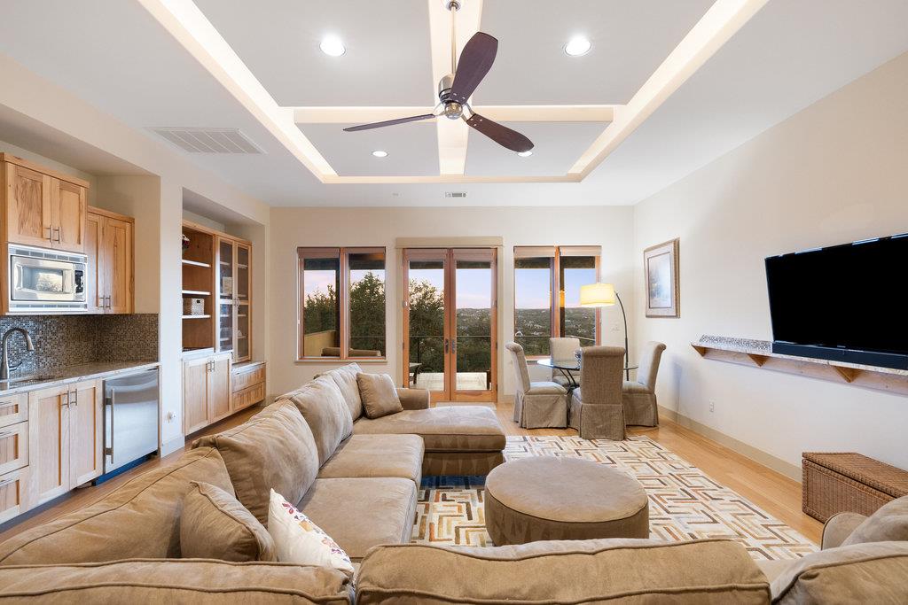 Open-plan living room and kitchen with a tan sectional, wood cabinetry and glass doors overlooking greenery.