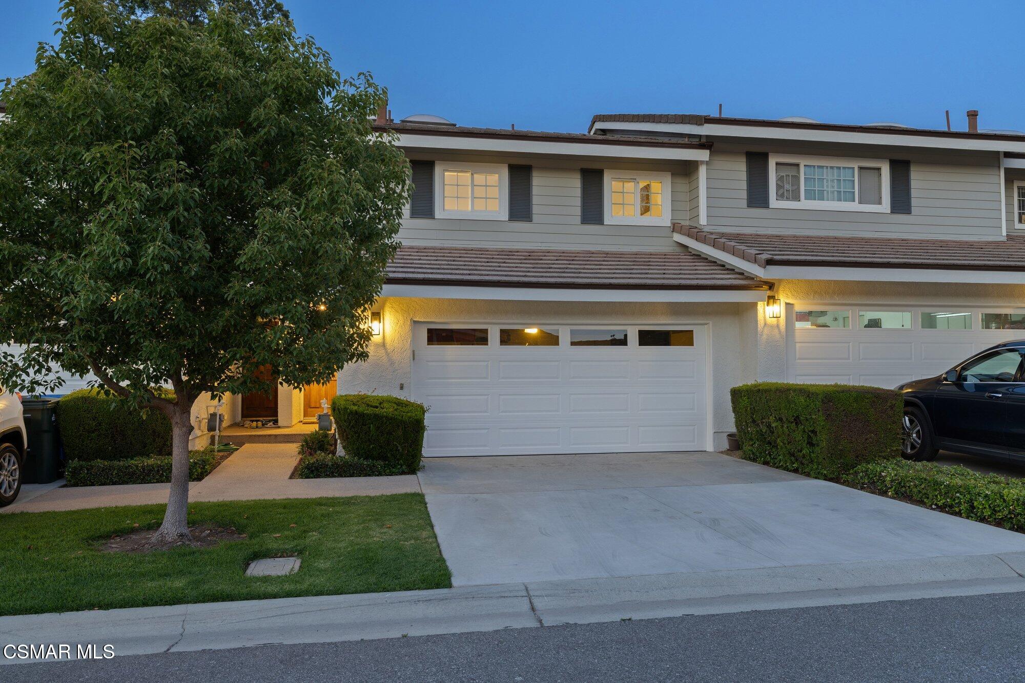 Two-story gray house at dusk with a large front tree, lit entry, and wide driveway.
