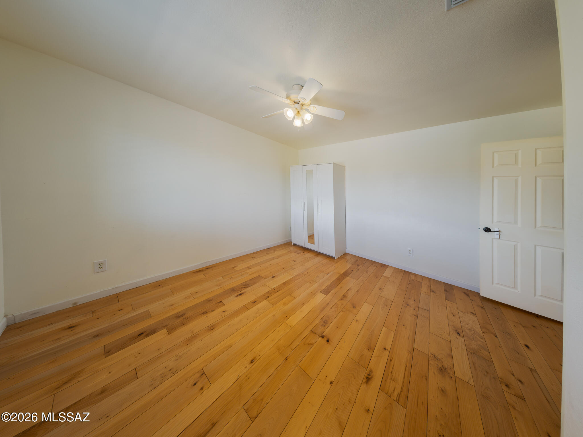 Empty bedroom with warm plank flooring, a mirrored wardrobe, and a lit ceiling fan.