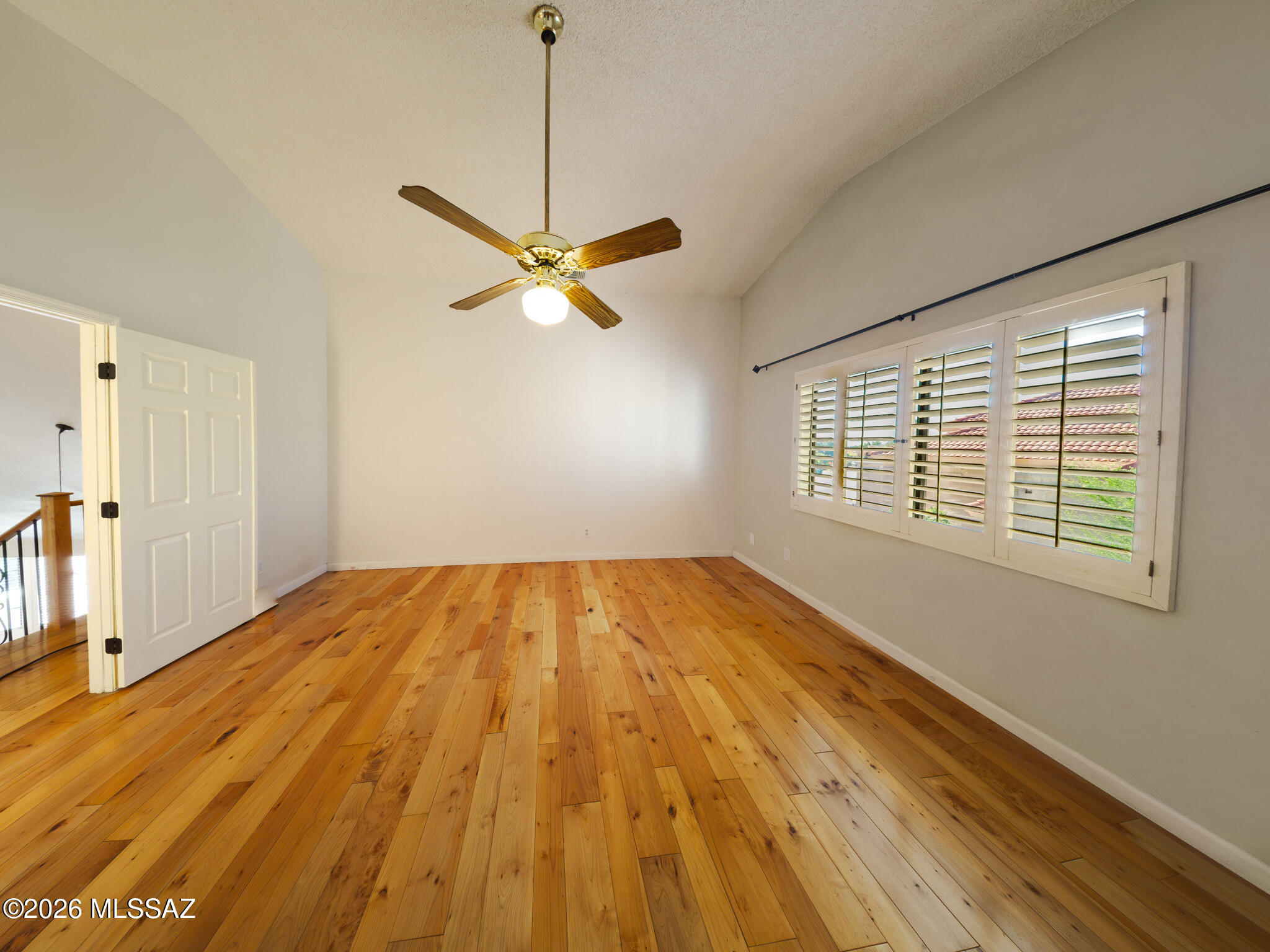 Empty room with hardwood floors, vaulted ceiling, ceiling fan, and plantation-shuttered windows.