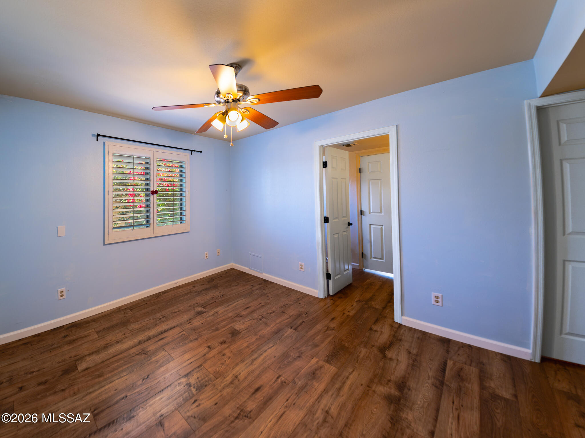 Empty room with blue walls, wood flooring, shuttered window, and illuminated ceiling fan.