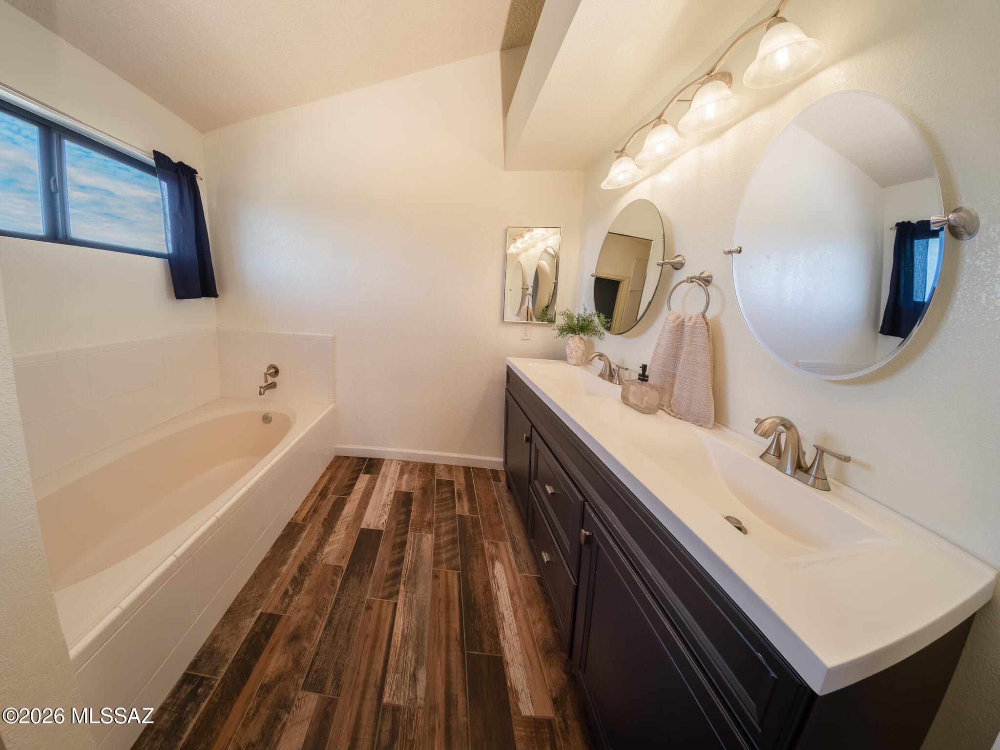 Bathroom with a white bathtub, double-sink dark vanity, large mirrors, and wood-look tile flooring.
