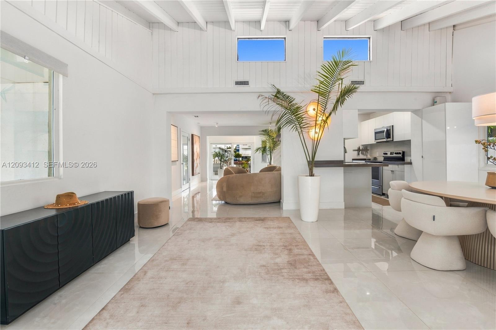 Open-plan living room with vaulted white ceiling, tall windows, and a potted palm beside the kitchen.
