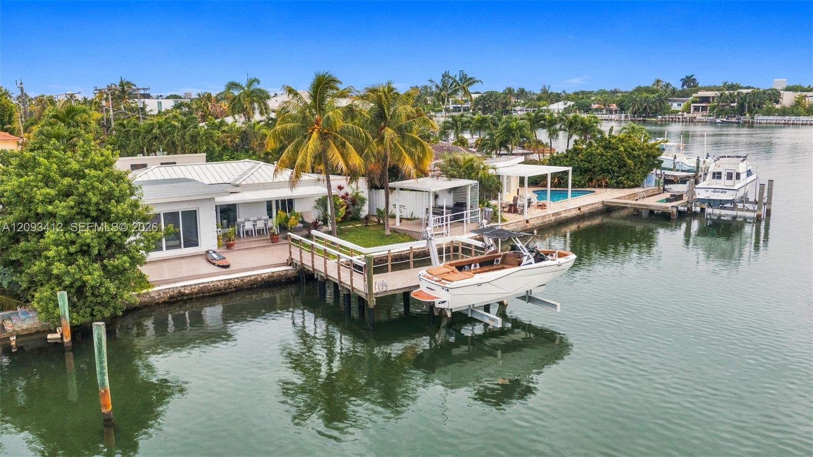 Aerial view of a waterfront home with a docked boat, pool, and palm-lined shoreline.