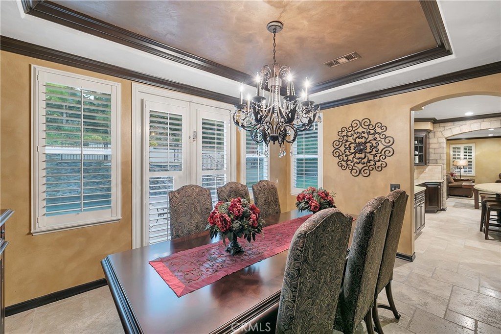 Dining room with an ornate chandelier, plantation shutters, and a dark wood table set with red flowers.