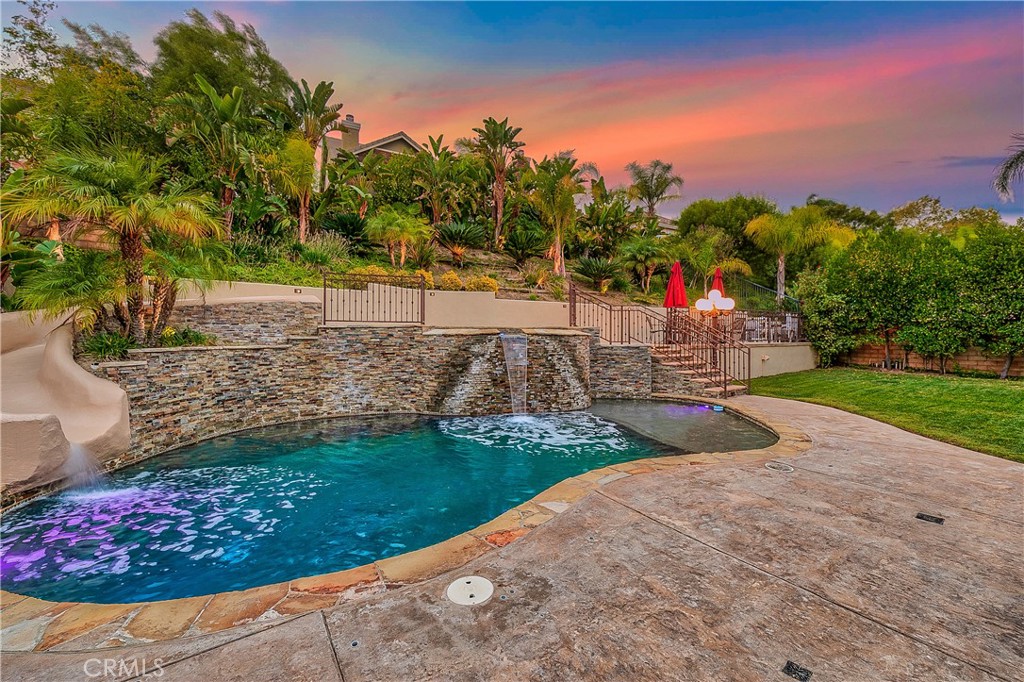 Backyard swimming pool with a stone waterfall, tropical landscaping, and a pink sunset.