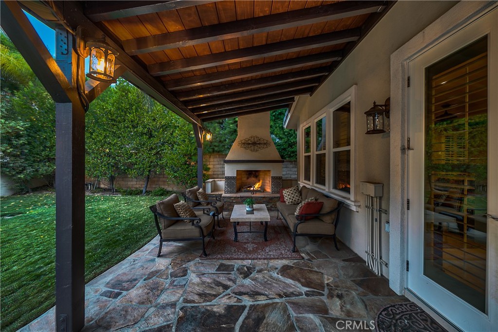 Covered stone patio with wood-beam ceiling, outdoor seating, and a lit fireplace overlooking the garden.