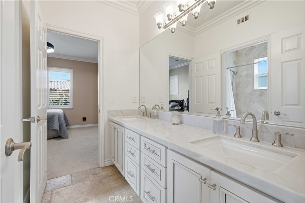 Primary bathroom with a long double-sink vanity and mirrored wall, opening to a carpeted bedroom.