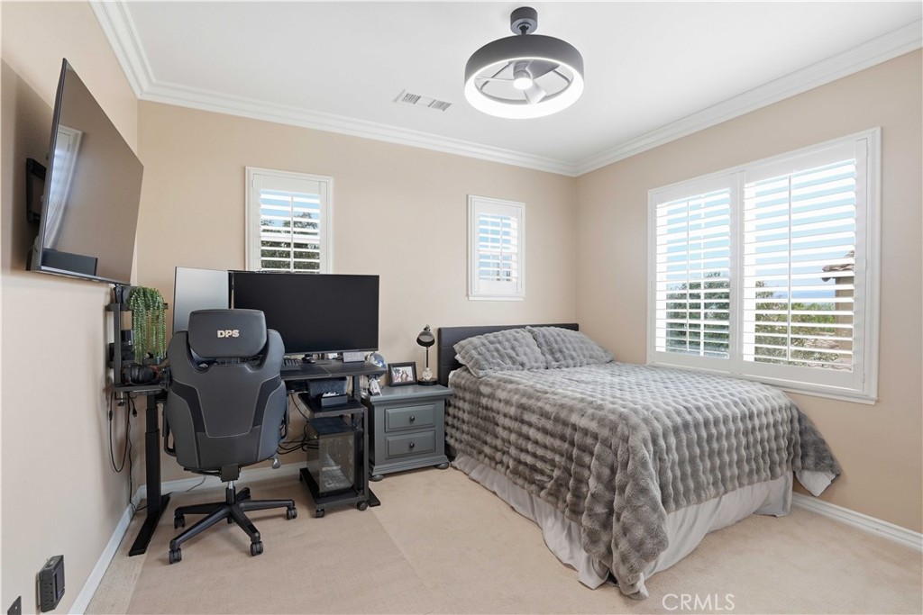 Bedroom with a gray textured bedspread, computer workstation, and wide windows with plantation shutters.