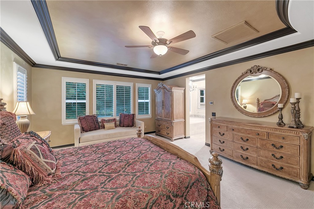 Bedroom with a patterned bed, window-seat sofa, carved dressers, and ornate oval mirror.
