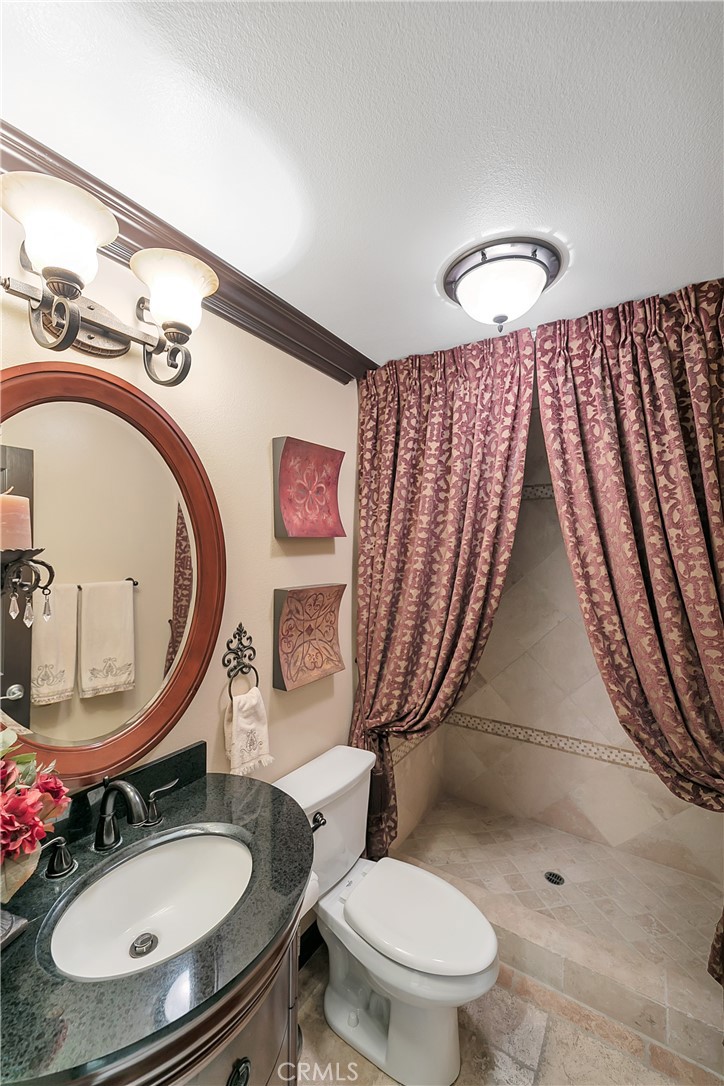 Bathroom with a dark stone vanity, oval mirror, toilet, and tiled shower behind patterned curtains.