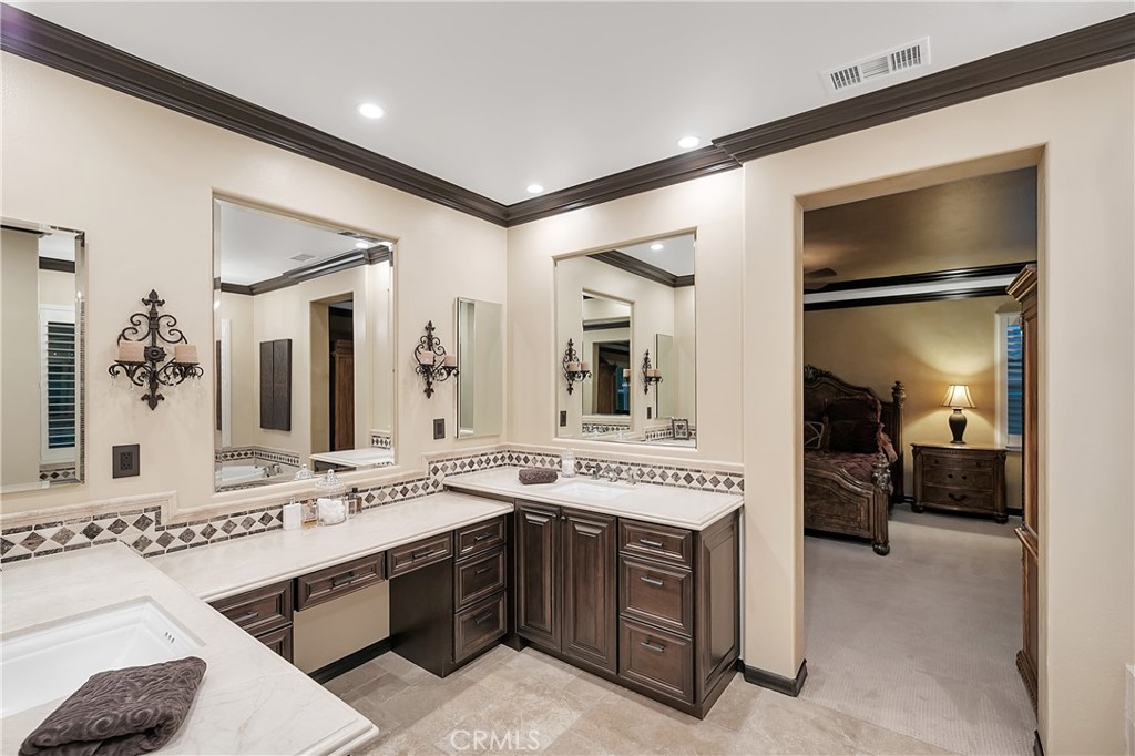 Bathroom with an L-shaped vanity, dark wood cabinets, and mirrors beside a furnished bedroom.