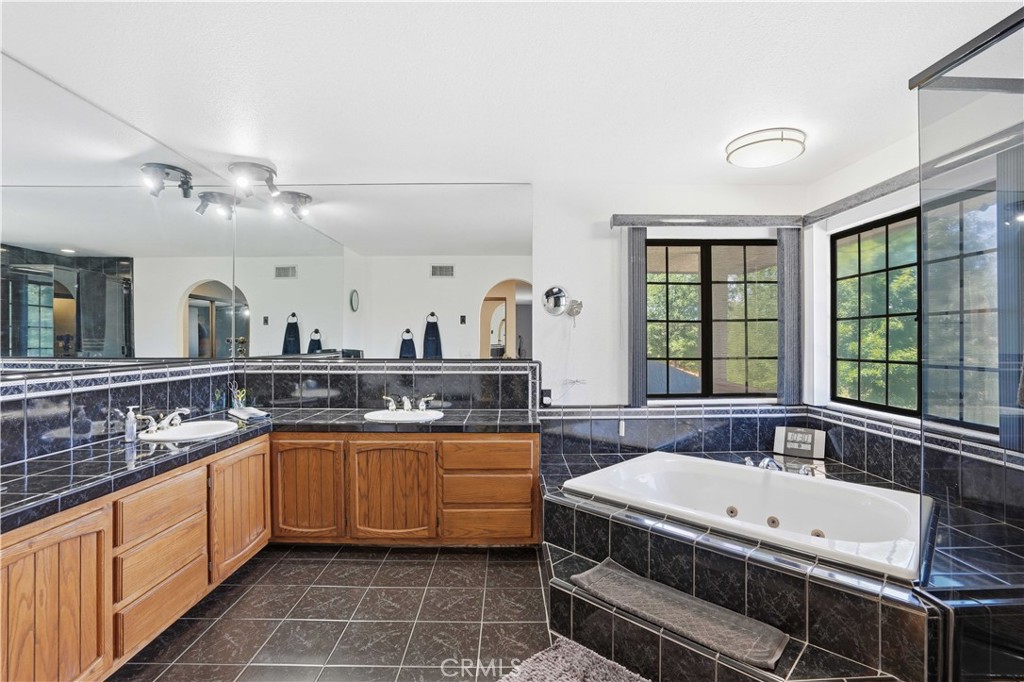 Spacious bathroom with a double-sink wood vanity, dark stone tile, and a jetted tub beside large windows.