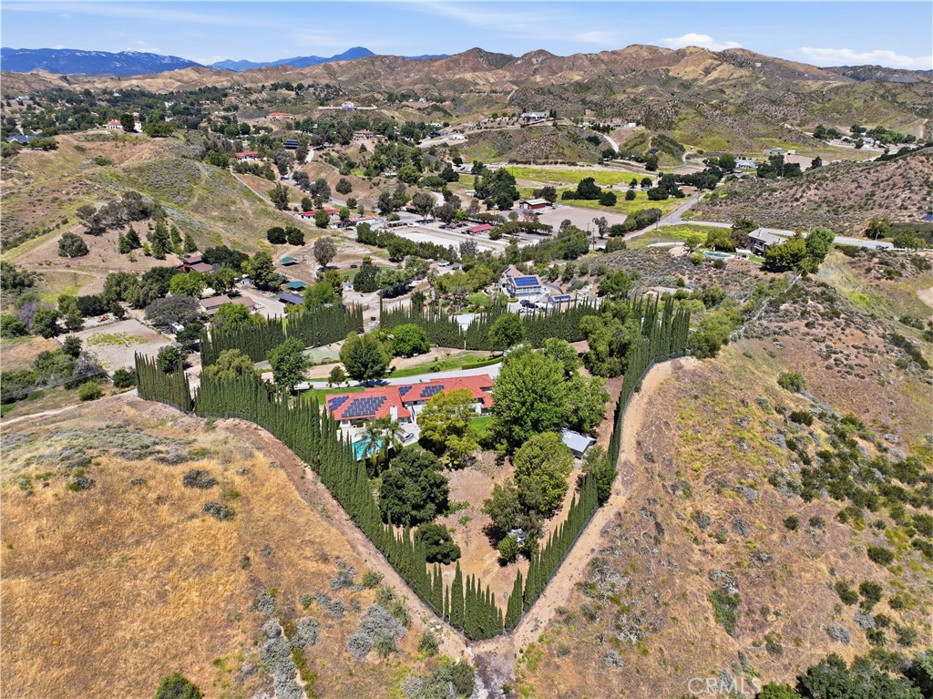 Aerial view of a hillside home with solar panels, a pool, and cypress trees, surrounded by dry hills.
