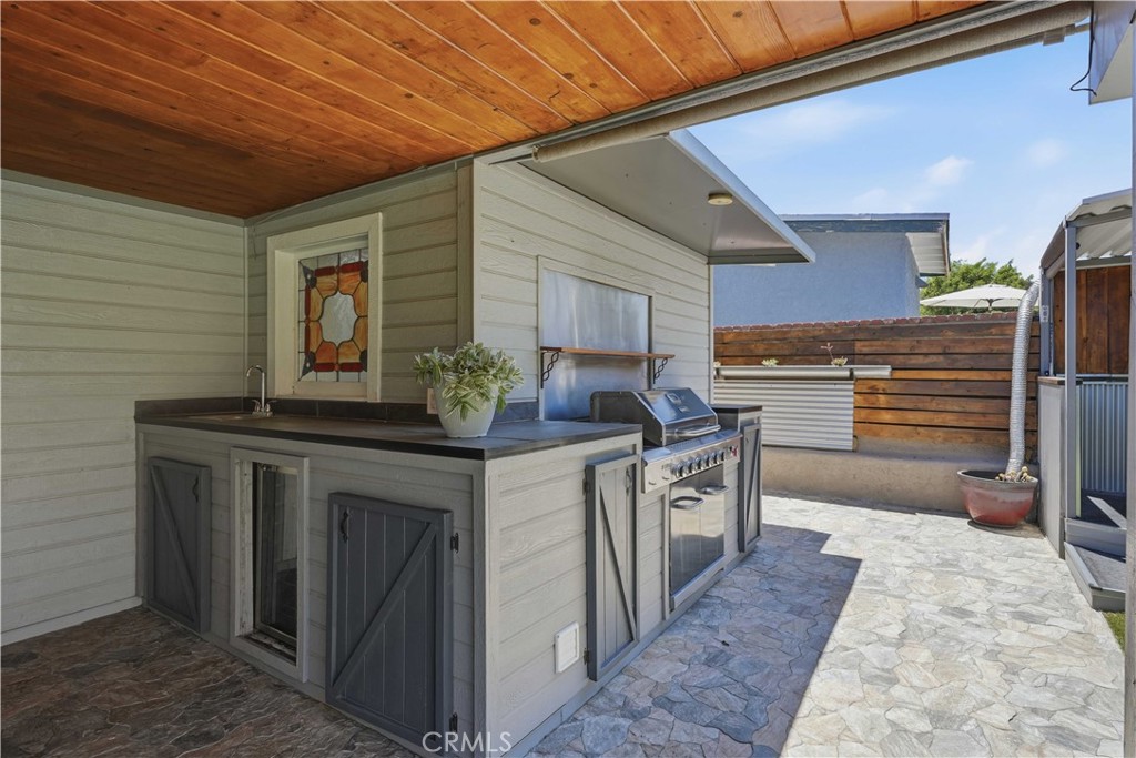 Covered patio kitchen with a built-in grill, sink, dark cabinets, and stone paving.