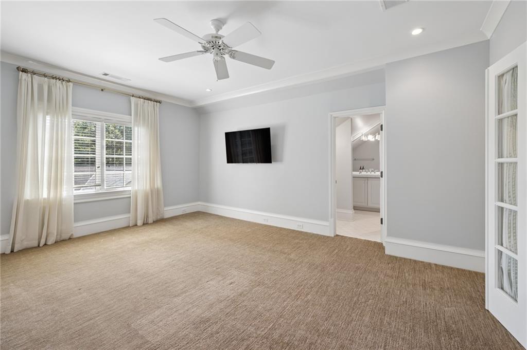 Empty bedroom with beige carpet, curtained windows, ceiling fan, and an open doorway to the bathroom.