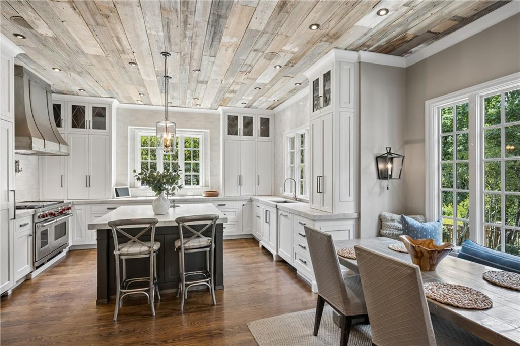 Kitchen with white cabinetry, dark island, reclaimed-wood ceiling, and adjoining dining area beside large windows.