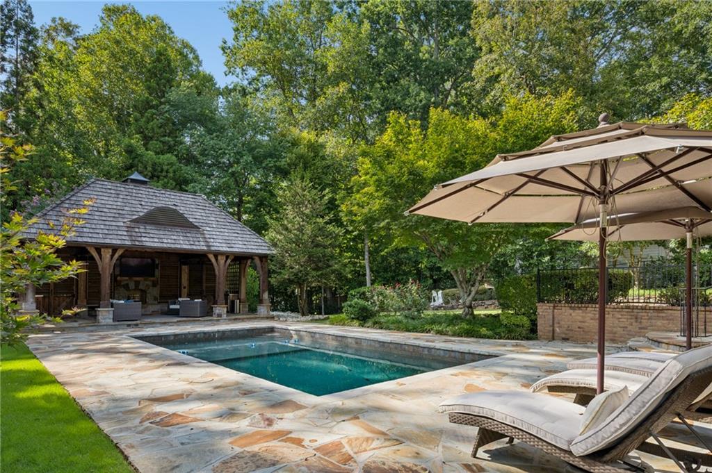 Backyard swimming pool beside a wooden pavilion, with stone patio, umbrellas, and lounge chairs.