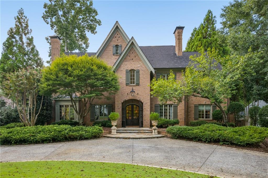 Brick home with steep gables, green shutters, mature landscaping, and a broad driveway.