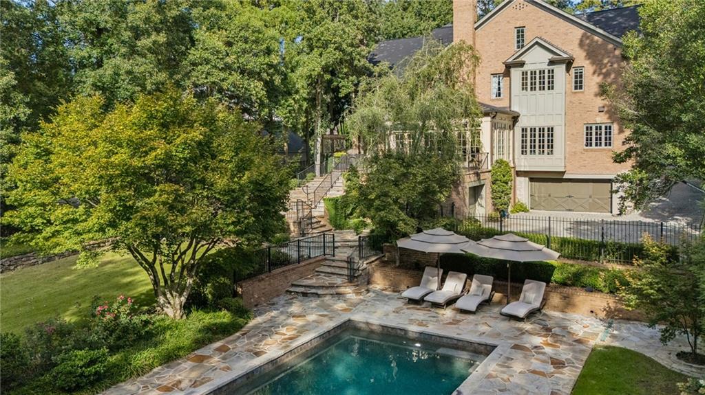 Brick home overlooking a stone pool terrace with lounge chairs, umbrellas, and mature trees.