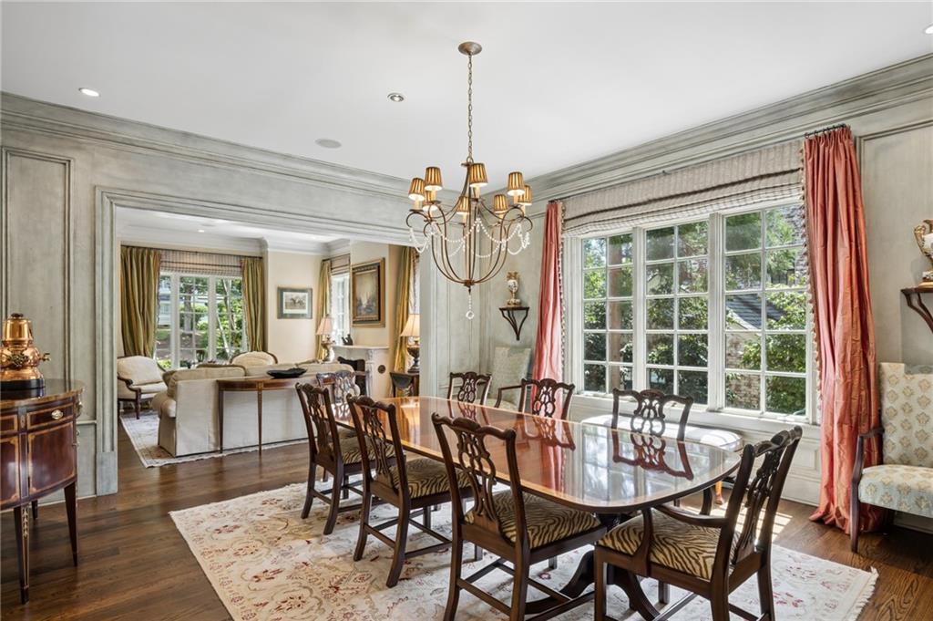 Dining room with a polished wood table, chandelier, and large window framed by coral curtains.