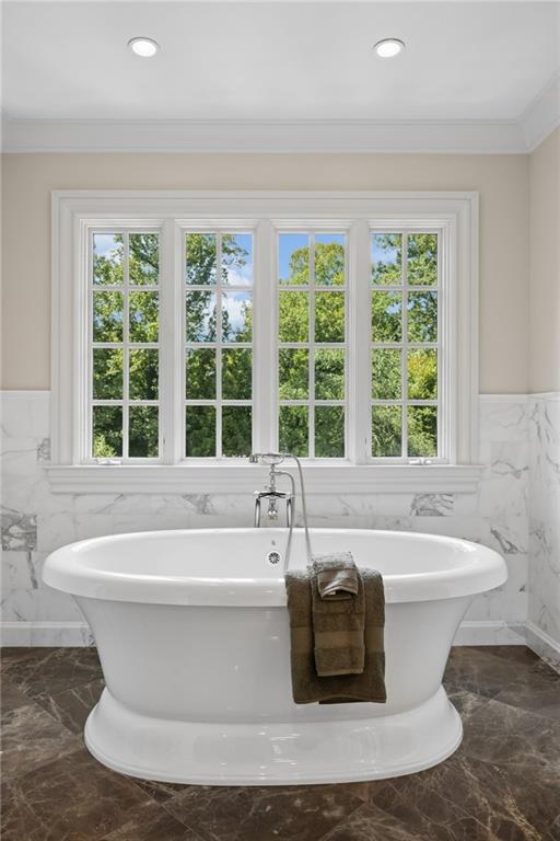 Freestanding soaking tub beneath a large window, with marble walls and a brown towel.
