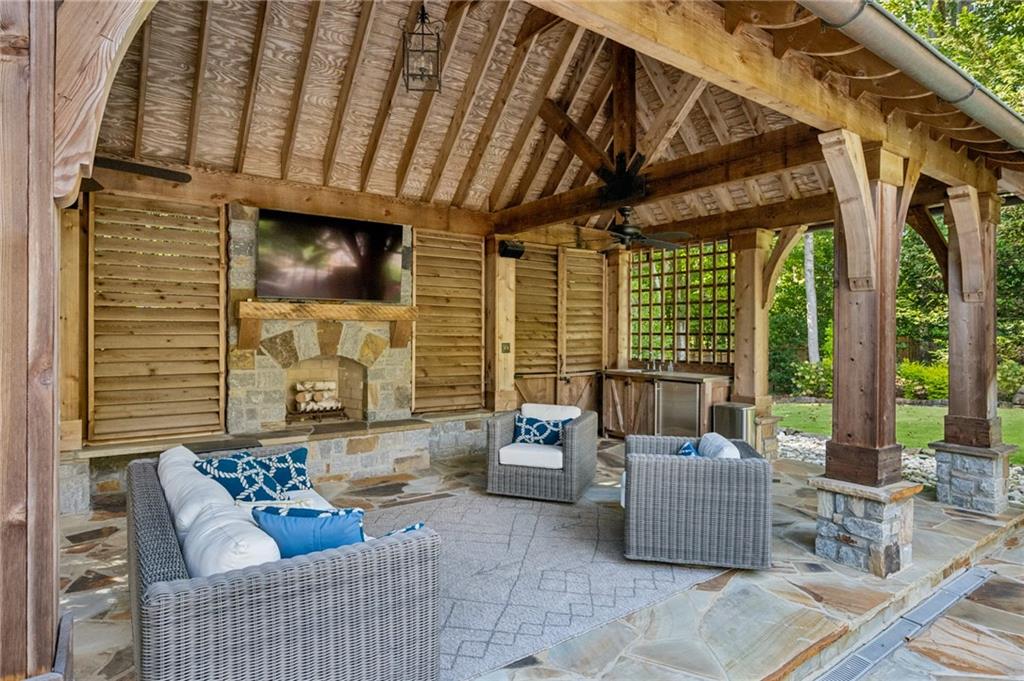 Covered patio with exposed timber beams, stone fireplace, and wicker seating overlooking a garden.