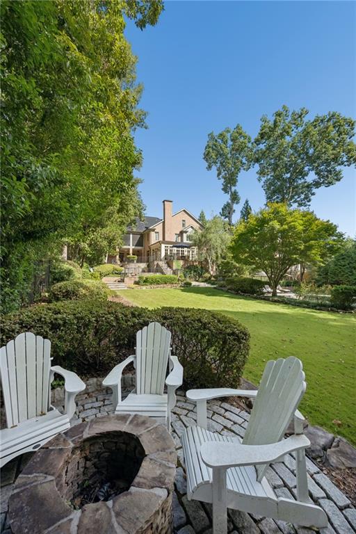 Backyard fire pit and white Adirondack chairs overlook a lawn and large brick house.