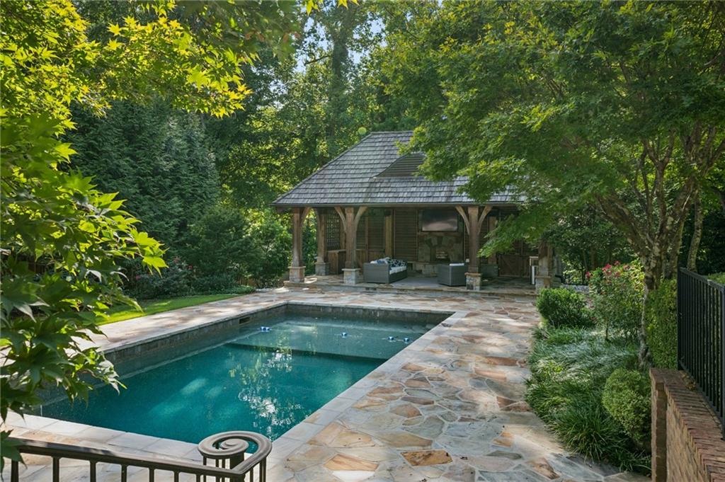 Swimming pool and stone patio beside a covered pavilion nestled among mature trees.