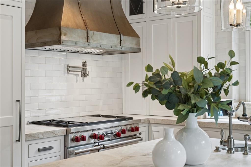 White kitchen with a stainless steel range, copper hood, subway tile backsplash, and greenery on the island.