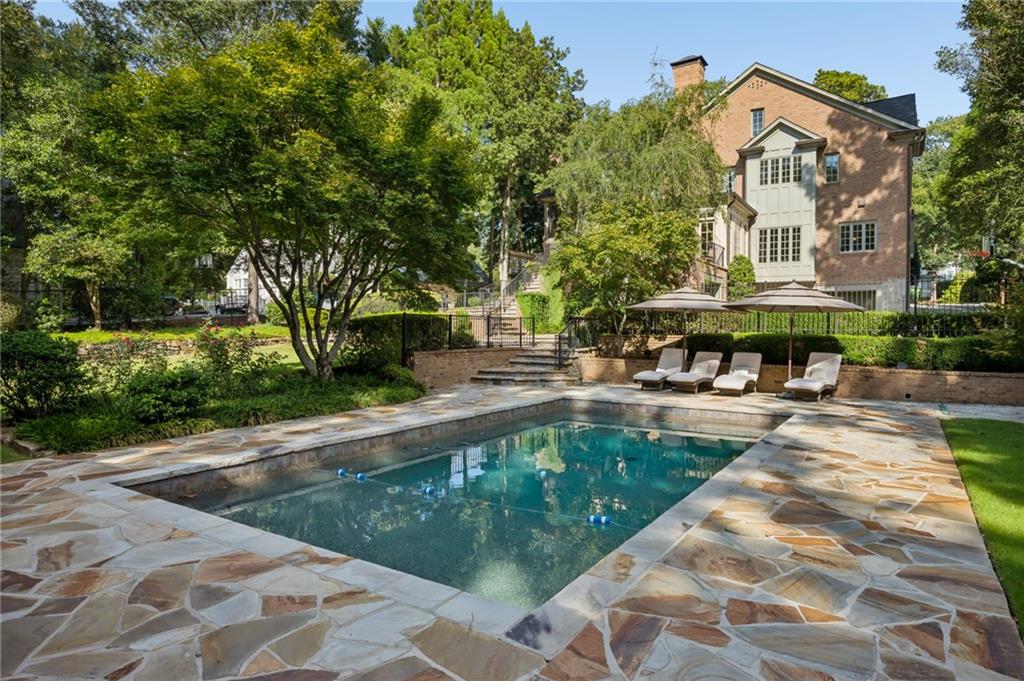 Backyard pool and stone patio beside a brick house, with landscaped greenery and umbrella-shaded loungers.
