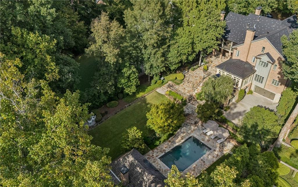 Aerial view of a brick home with a swimming pool, stone patios, and a tree-lined lawn.