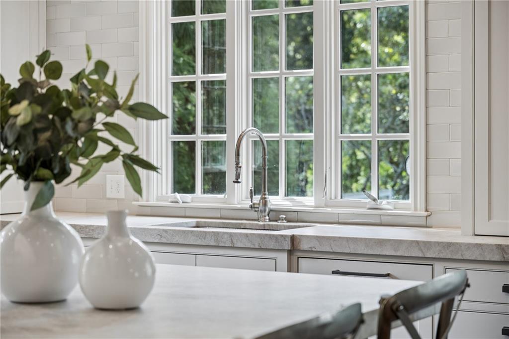 Kitchen sink beneath tall divided-light windows, with a white tile backsplash and decorative vases on the island.