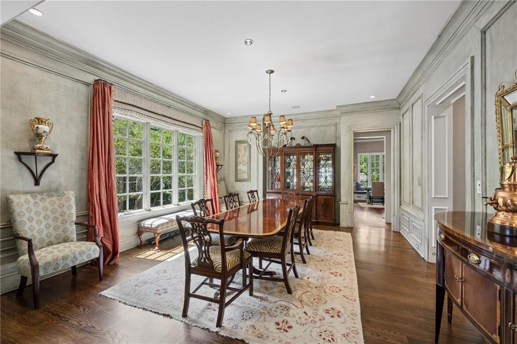 Dining room with a polished wood table, chandelier, paneled walls, and large curtained windows.