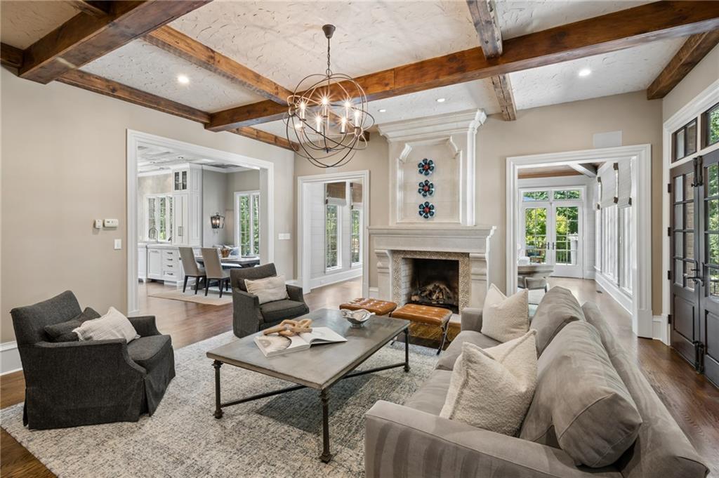 Living room with exposed wood beams, a stone fireplace, and tall windows overlooking greenery.