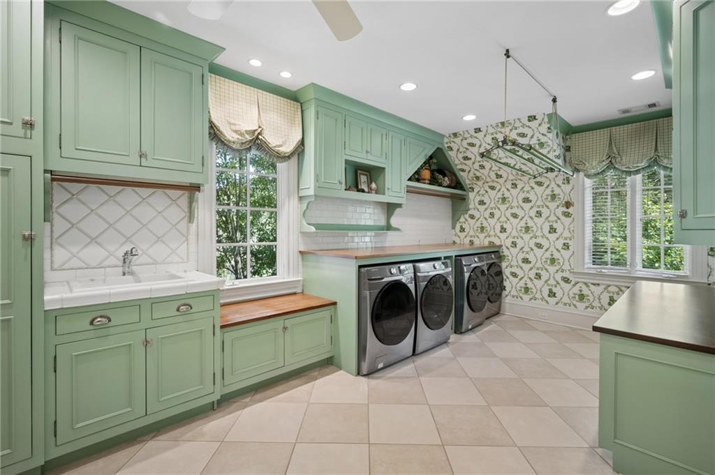 Laundry room with green cabinetry, three front-loading machines, a deep sink, and patterned wallpaper.