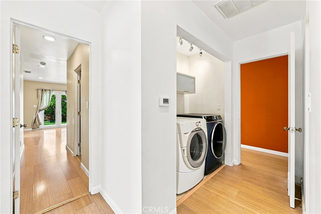 Laundry area with front-loading washer and dryer beside a hallway and orange-walled room.