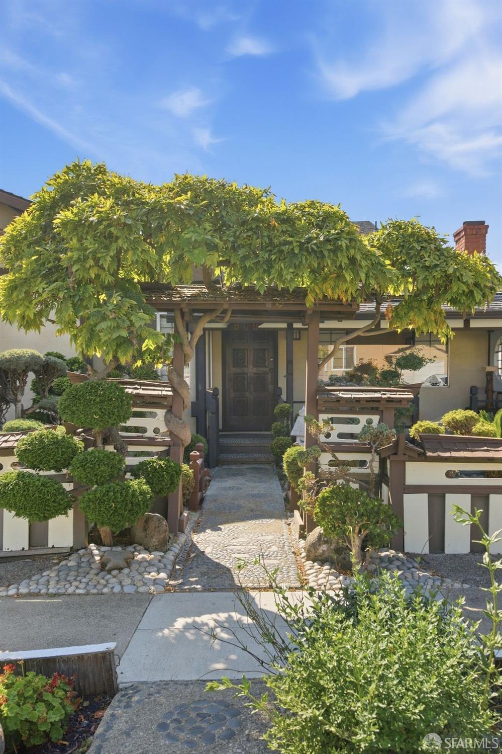 Home entrance beneath a leafy tree, approached by a stone path through a landscaped garden.