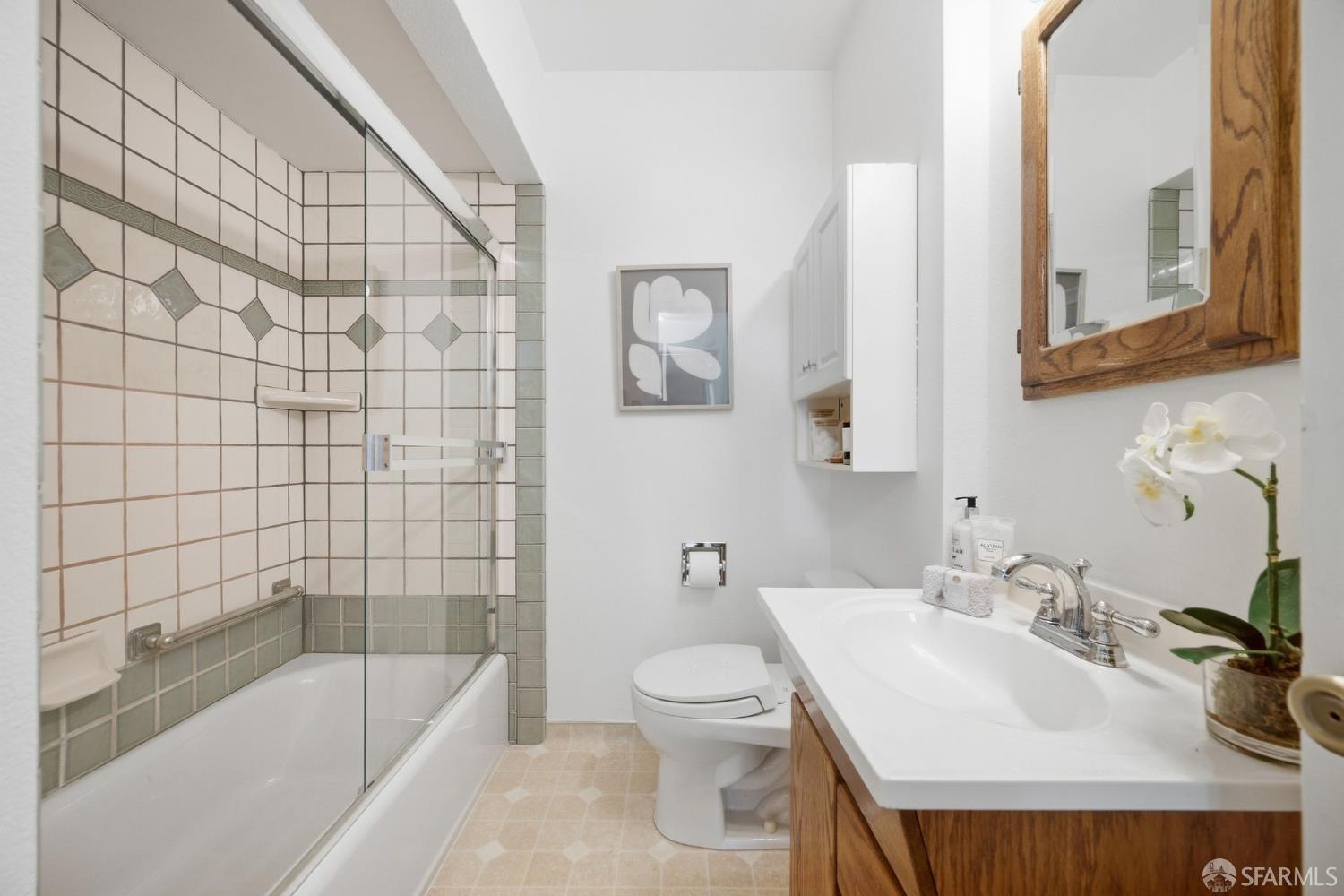 Bathroom with a glass-enclosed tiled tub and shower, wood vanity, and orchid beside the sink.