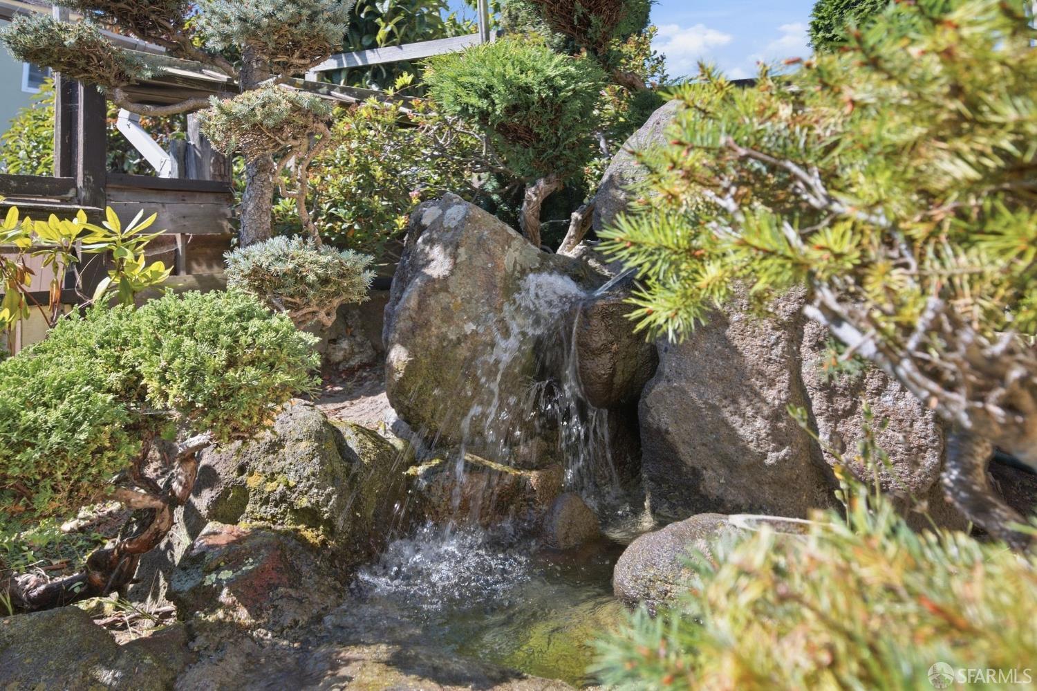 Rock waterfall flowing into a garden pond among dense shrubs and trees.