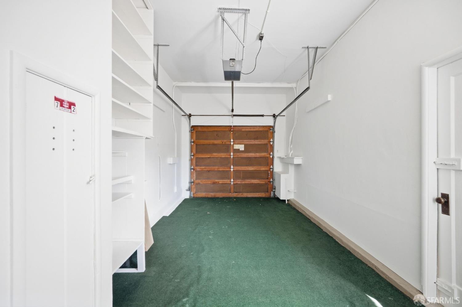 Empty garage with green carpet, built-in shelving, and a wooden overhead door.