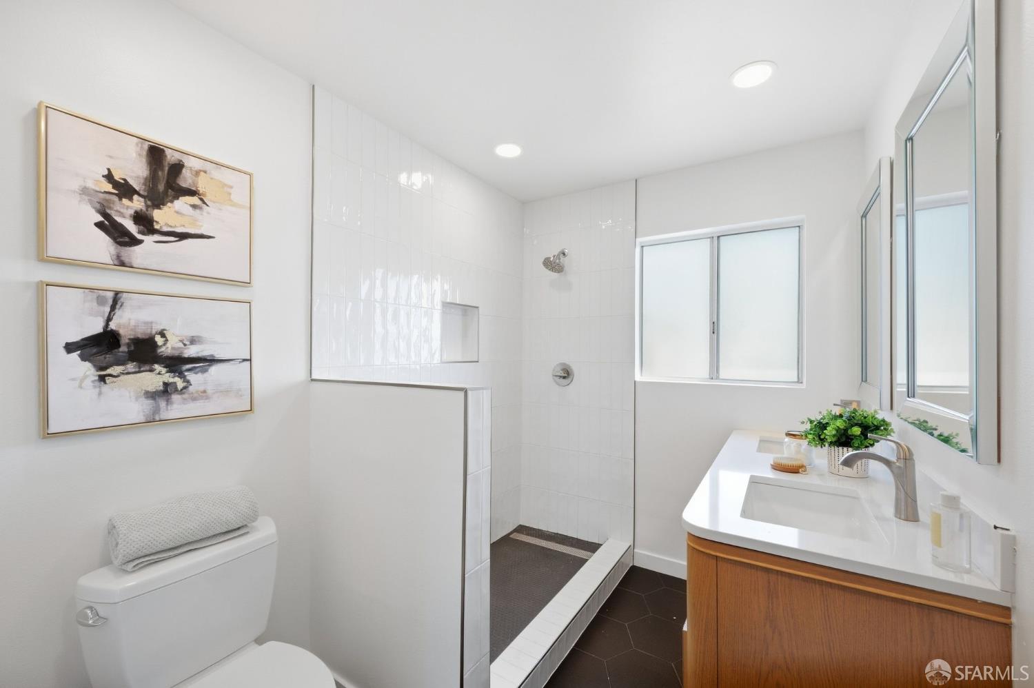 Bathroom with a white tiled walk-in shower, wood vanity, and abstract paintings above the toilet.