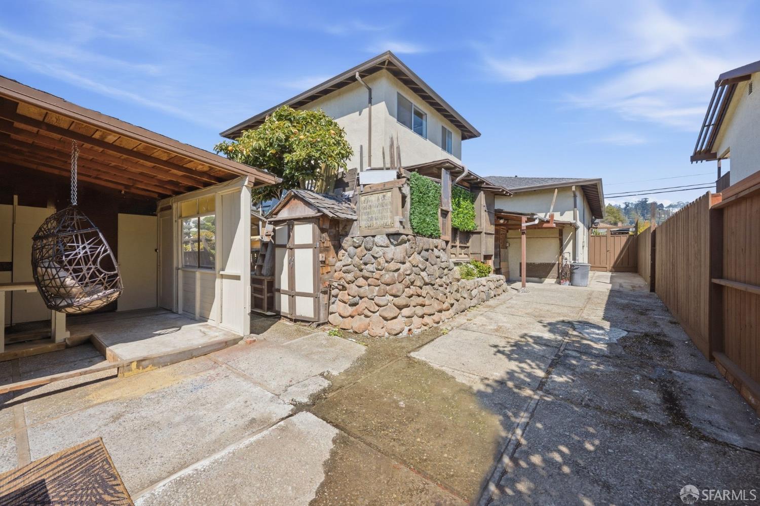 Home’s rear courtyard with a covered patio, hanging chair, stone retaining wall, and fenced driveway.
