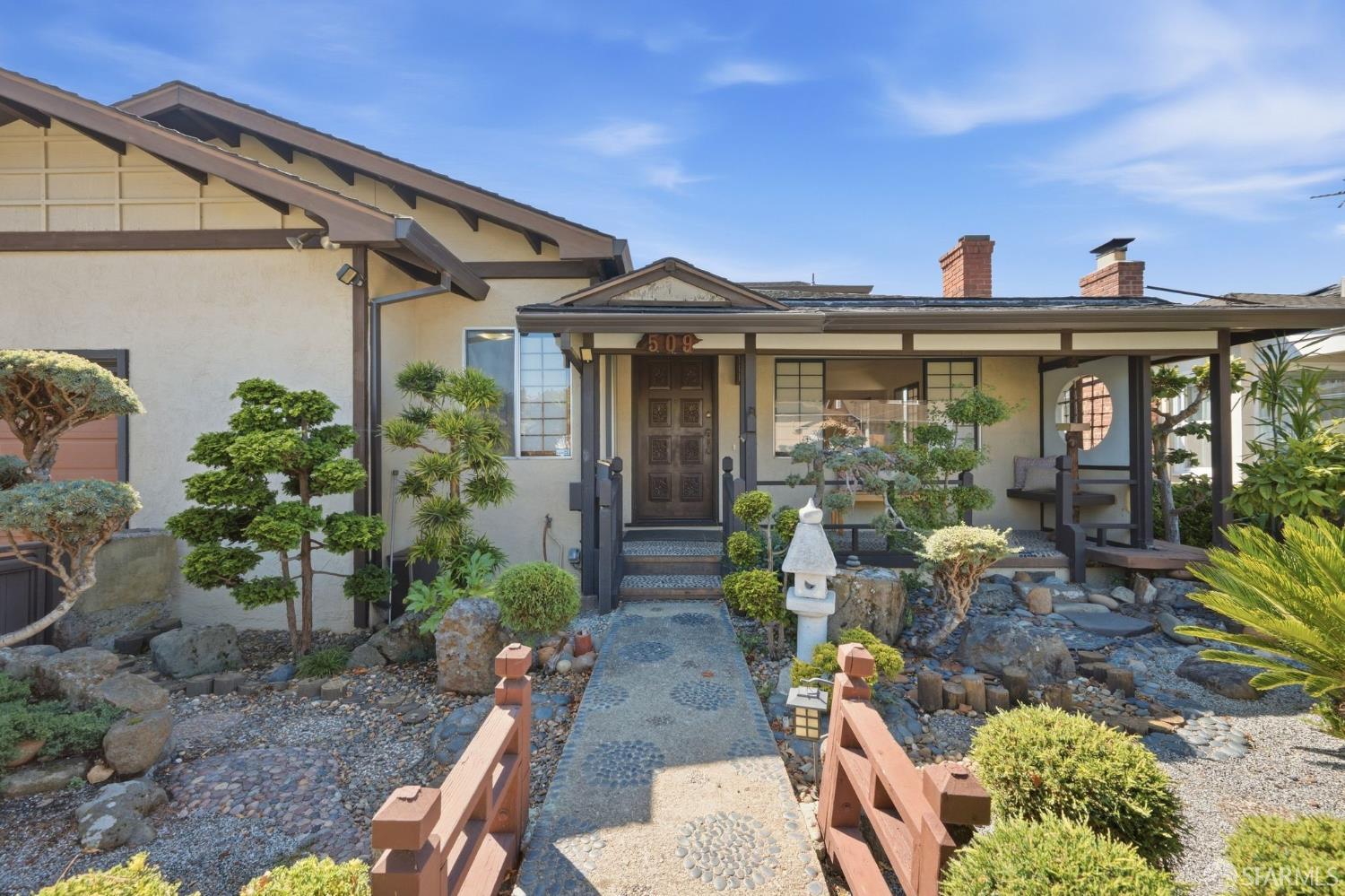 Home exterior with covered entry, landscaped rock garden, and pebble walkway over a small wooden bridge.